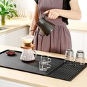 Zwarte mat met een vrouw die koffie zet met een filter, een glas, twee kleine kopjes, drie glazen op een afdruiprek en koffiebonen.
