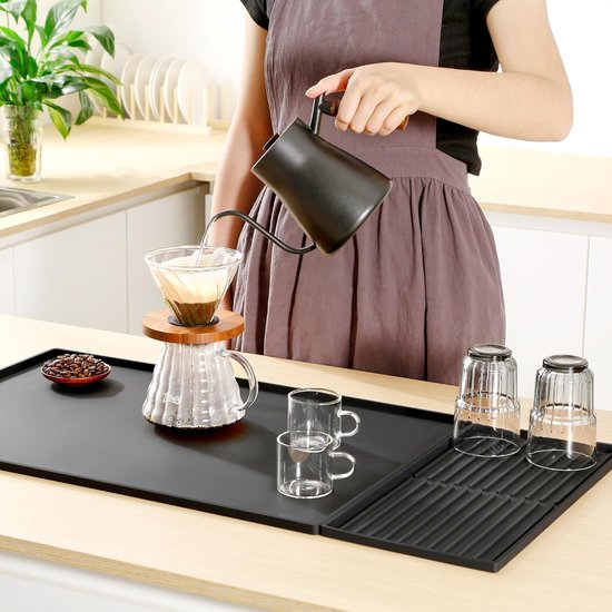 Zwarte mat met een vrouw die koffie zet met een filter, een glas, twee kleine kopjes, drie glazen op een afdruiprek en koffiebonen.