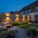 Houten schutting met vier zwarte, kubusvormige wandlampen die naar boven en beneden schijnen. Naast een loungehoek in de tuin.
