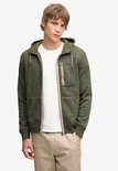 Model draagt olijfgroene, gemêleerde fleece hoodie met beige rits, zijzak en Superdry & Co-badge.