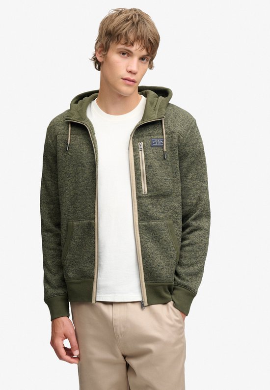 Model draagt olijfgroene, gemêleerde fleece hoodie met beige rits, zijzak en Superdry & Co-badge.
