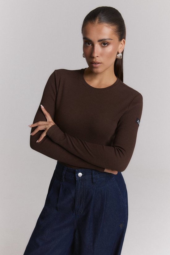 Model draagt bruine gebreide longsleeve top met logo op de mouw, gecombineerd met jeans.