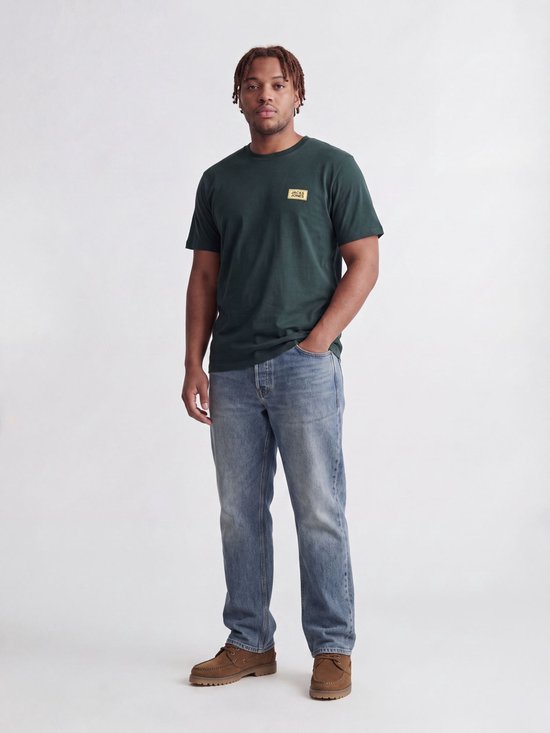 Man met gevlochten haar draagt een donkergroen T-shirt met geel logo op de borst en een spijkerbroek.
