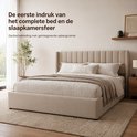 Een beige gestoffeerd tweepersoonsbed met een hoge, gestreepte hoofdsteun en geïntegreerde opbergruimte, in een lichte slaapkamer met planten en een raam.