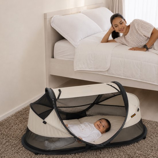 Een vrouw kijkt toe hoe een baby in een wit en zwart Deryan campingbedje ligt, naast een bed.