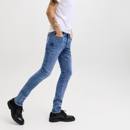 Man in een witte T-shirt en een blauwe spijkerbroek met zijn handen in zijn zakken, lopend met zwarte schoenen.