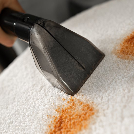 Een grijze reinigingsmondstuk zuigt oranje vlekken op van een witte handdoek of badstof.