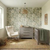 Babykamer met grijs houten babybed, commode en een schommelstoel tegen een behang met bladeren en luipaarden.