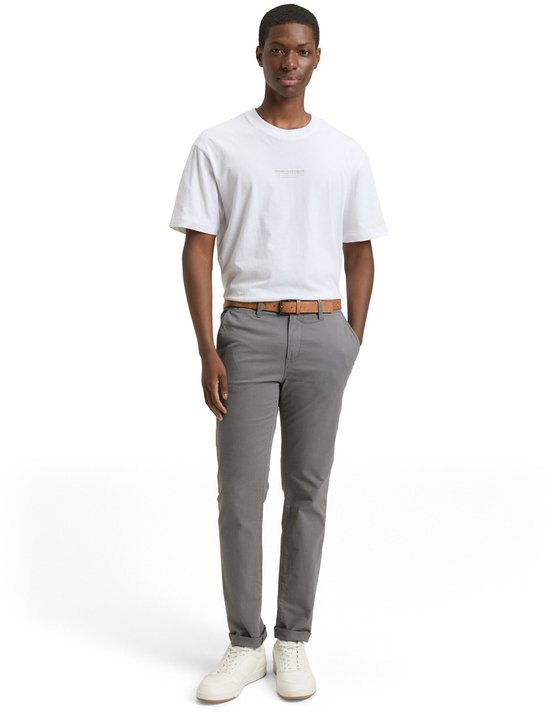 Man in grijze chino met bruine riem, wit T-shirt en witte sneakers.
