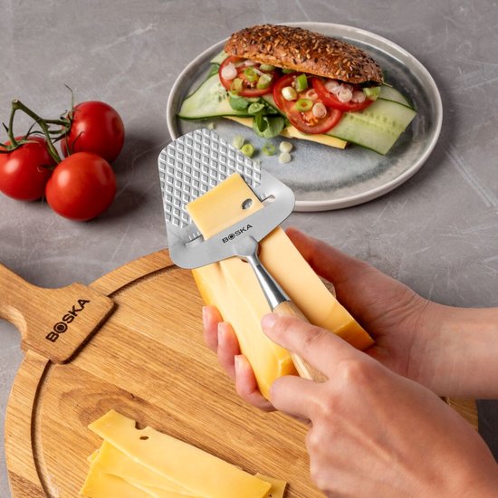 Handen gebruiken een kaasrasp met houten handvat om een stuk kaas te snijden, boven een houten plank.