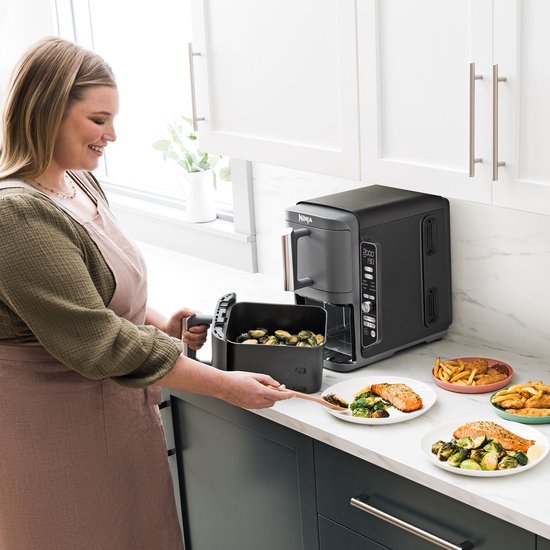 Vrouw met schort die groenten uit een zwarte Ninja heteluchtfriteuse haalt, naast zalm en friet.