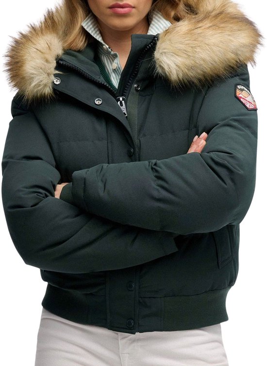 Gedetailleerde opname van een vrouw in een donkergroene bomberjack met bontkraag en logo patch op de mouw. Ze draagt een gestreepte blouse en kabeltrui, armen gekruist.