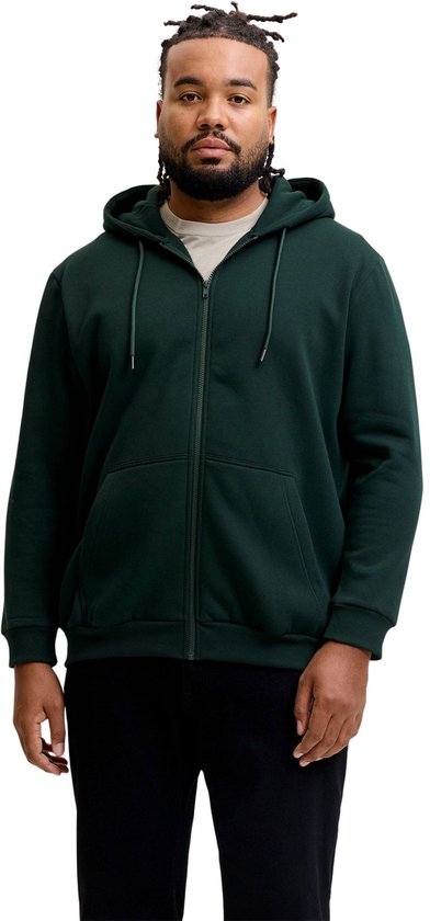 Man met dreadlocks, donkergroene hoodie, beige T-shirt en zwarte broek, staand met handen in zakken.