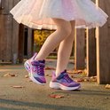 Paarse Skechers sneakers met regenboog-accenten, roze veters en velcro sluiting. Op een meisje met beige maillot en tutu.