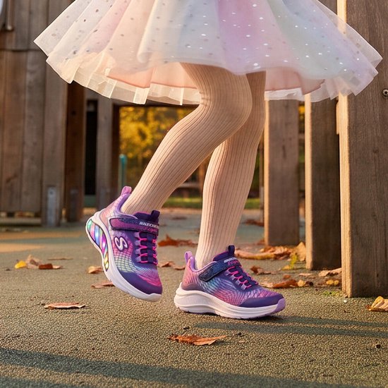 Paarse Skechers sneakers met regenboog-accenten, roze veters en velcro sluiting. Op een meisje met beige maillot en tutu.