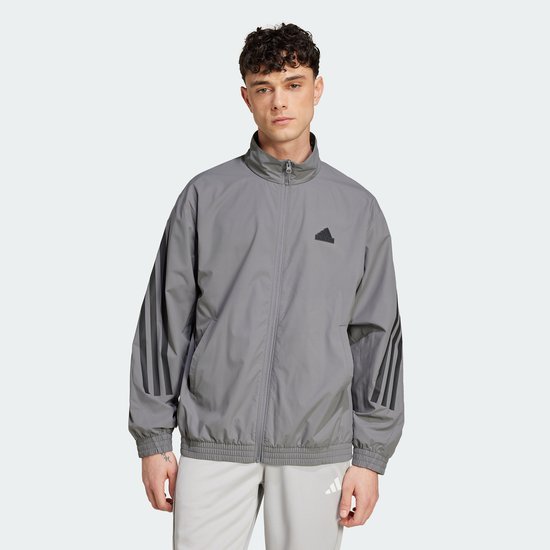Man draagt een grijs Adidas trainingsjack met zwarte 3-strepen op de mouwen en een Adidas logo op de borst.