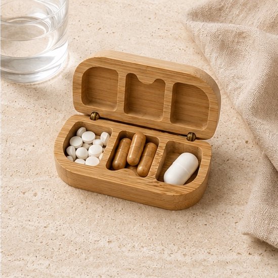 Open houten pillendoosje met 6 vakjes, gevuld met diverse pillen en capsules, naast een glas water.