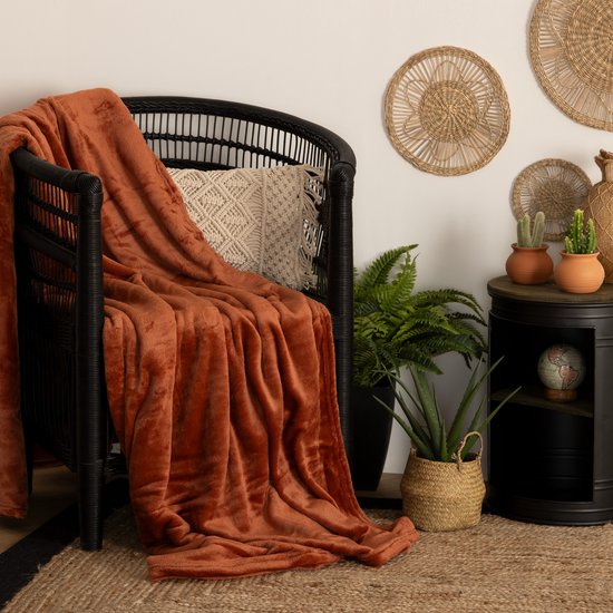 Terracotta fleece deken gedrapeerd over een zwarte rotan stoel, met kussens en planten.