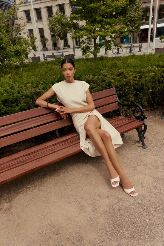 Een model met donker haar zit op een houten bankje in een park. Ze draagt een crème-kleurige jurk en witte sandalen.