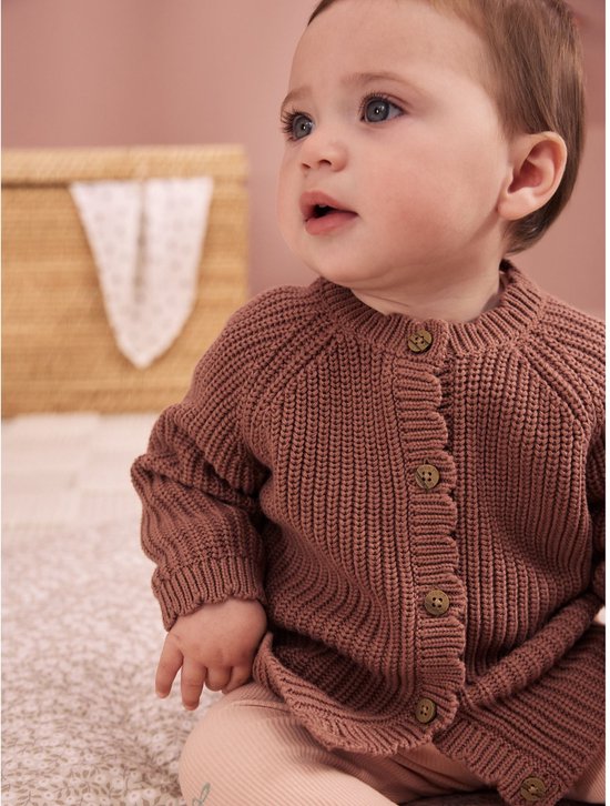 Baby zit op de grond in een bruin gebreid vest met knopen.