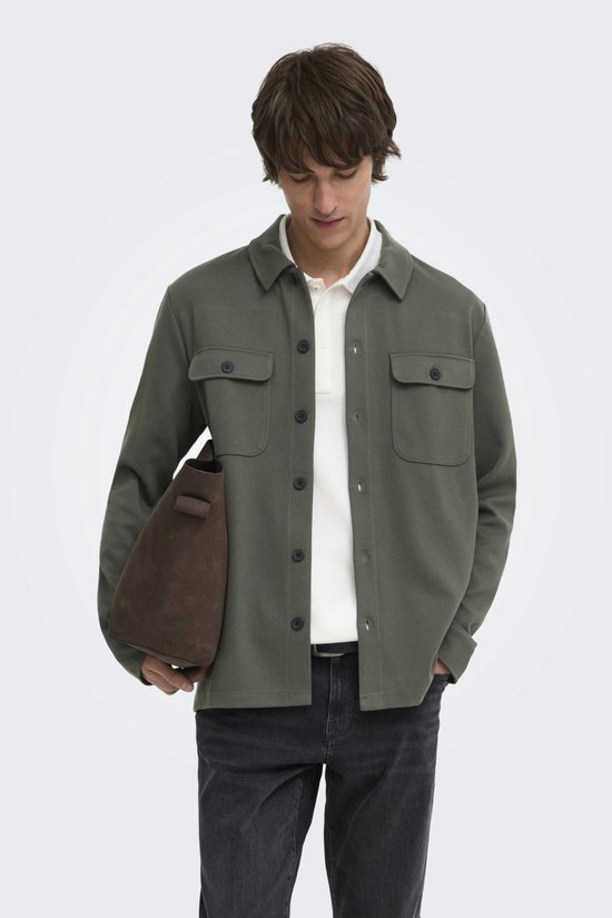 Mannelijk model met olijfgroene, lange mouwen oversized shirt, witte polo eronder en grijze jeans.