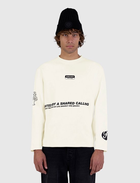 Model met zwarte muts draagt een ecru longsleeve met 'Pure Path' tekst op de borst en zwarte broek.