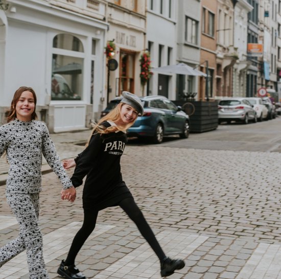 Twee meisjes lopen over een zebrapad in een stad. Het meisje rechts draagt een zwart jurkje met witte kraag en 'PARIS CHAMP DE MARS' opdruk.
