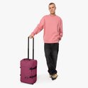 Man met roze trui en zwarte broek staat naast bordeauxrode koffer met zwarte banden.