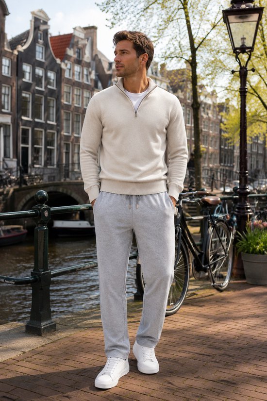 Man met grijze joggingbroek en beige trui met ritskraag, witte sneakers aan gracht.