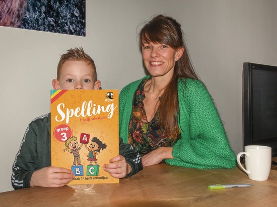 Een jongen houdt een "Spelling Groep 3" oefenboek omhoog, een vrouw kijkt lachend toe. Het boek toont twee kinderen met de letters A, B, C.