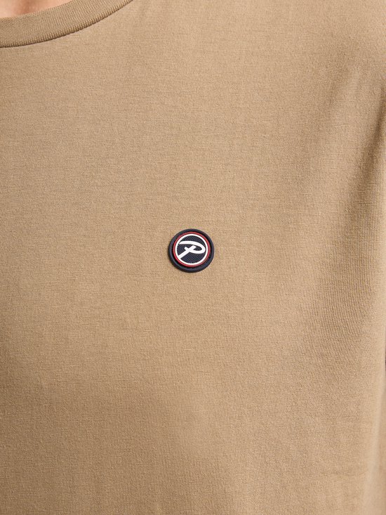 Close-up van een geborduurd logo in rood, wit en blauw op een lichtbruin t-shirt.