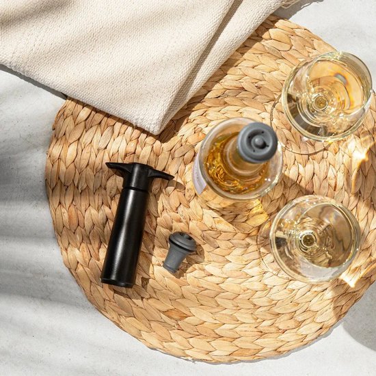 Zwarte Vacu Vin wijnstopper met een fles witte wijn en twee glazen op een rieten onderzetter.