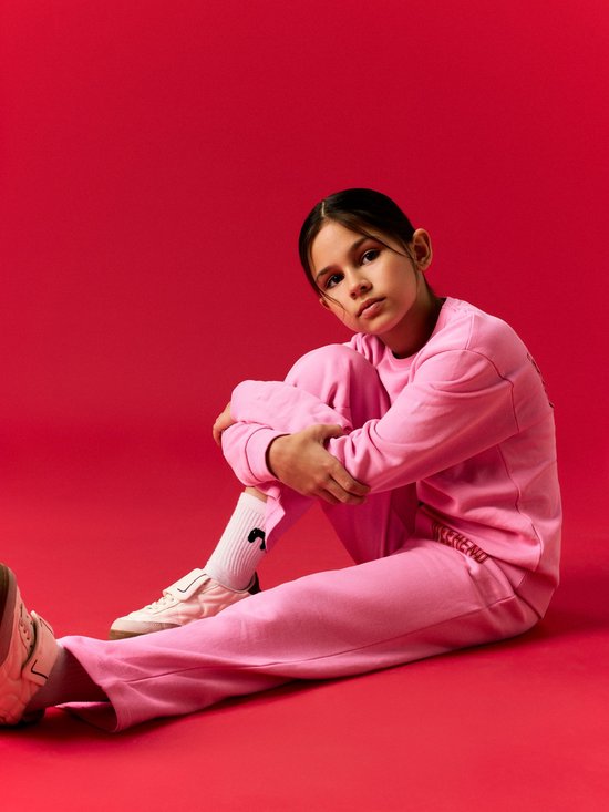 Meisje in een roze joggingbroek en sweater. De tekst 'WEEKEND' is zichtbaar op de broek.