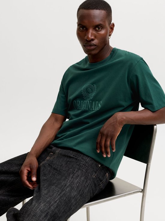 Man zittend in een donkergroen Jack & Jones T-shirt met "ORIGINALS" opdruk.