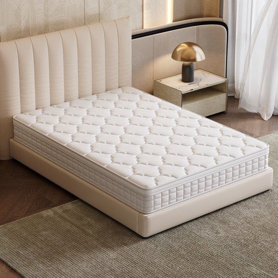 Een witte matras met gebreid patroon staat op een bed in een slaapkamer met een beige hoofdbord.