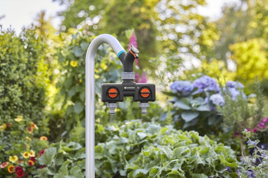 Zwarte Gardena waterverdeler met twee oranje draaiknoppen, aangesloten op een kraan in een tuin met bloemen.