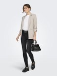 Model in een beige blazer met opgerolde mouwen, witte top, zwarte skinny jeans, riem en zwarte tas.