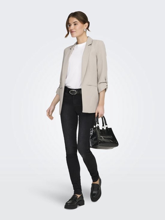 Model in een beige blazer met opgerolde mouwen, witte top, zwarte skinny jeans, riem en zwarte tas.