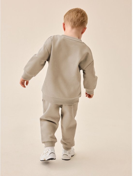 Rugzijde van een jongen in een naturelkleurige sweater en joggingbroek met witte sneakers.