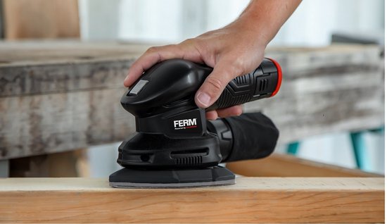 Hand houdt een zwarte FERM palm schuurmachine vast boven een houten plank.