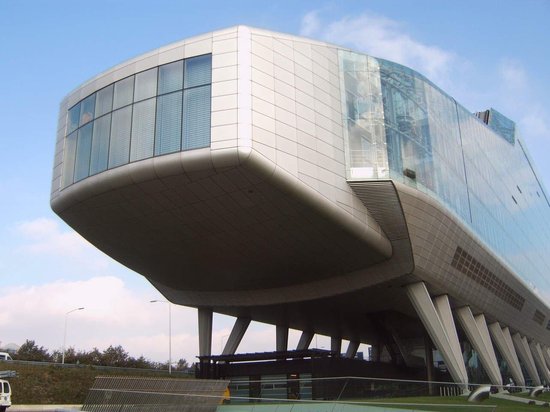 Modern, futuristisch gebouw op palen met een glazen gevel en een gebogen, metalen dak. Deels gespiegelde ramen.