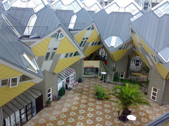 Een complex van grijze en gele kubuswoningen met schuine daken en veel ramen, rond een betegeld plein met een palm.