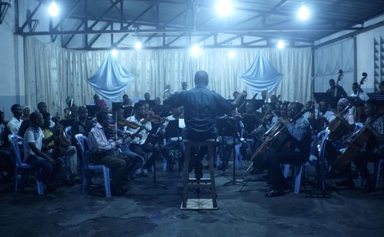 Een orkest speelt in een kamer met blauwe draperieën aan de muur. Een dirigent staat met zijn rug naar de camera.