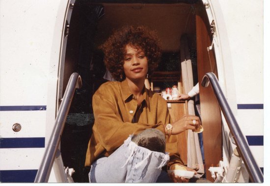 Whitney Houston in een okergele blouse en jeans zit op de trap van een vliegtuig.
