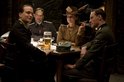 Vier personen aan een tafel met bier en sigarettenrook, waarbij drie mannen militaire uniformen dragen en een vrouw een hoed.