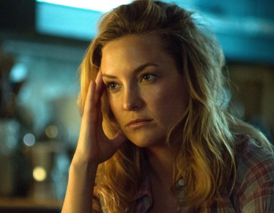Kate Hudson met blond haar, leunend met haar hand tegen haar hoofd, draagt een geruit shirt.