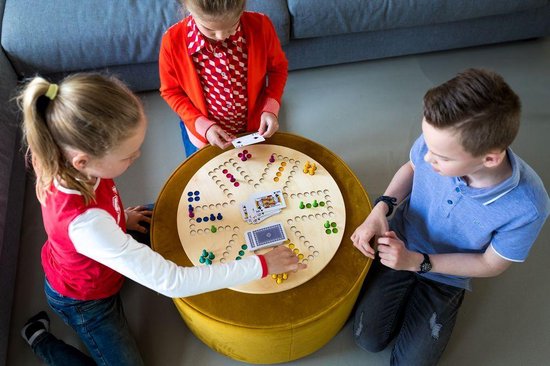 Drie kinderen spelen een bordspel, gezien van bovenaf. Het spel heeft gekleurde pionnen en speelkaarten op een rond houten bord.