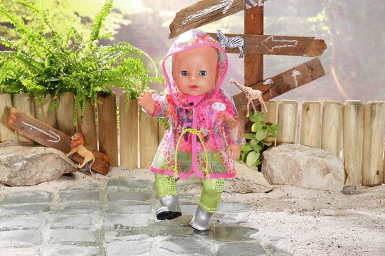Baby pop draagt roze shirt met 'BE WILD', groene broek, zilveren laarsjes en een doorzichtige regenjas met regenboogprint.