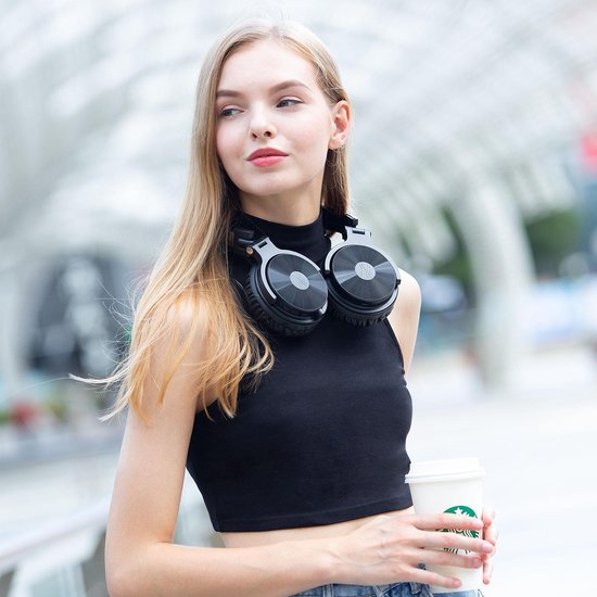 Vrouw met blonde haren in een zwart crop top, draagt een zwarte koptelefoon om haar nek en houdt een koffiebeker vast.