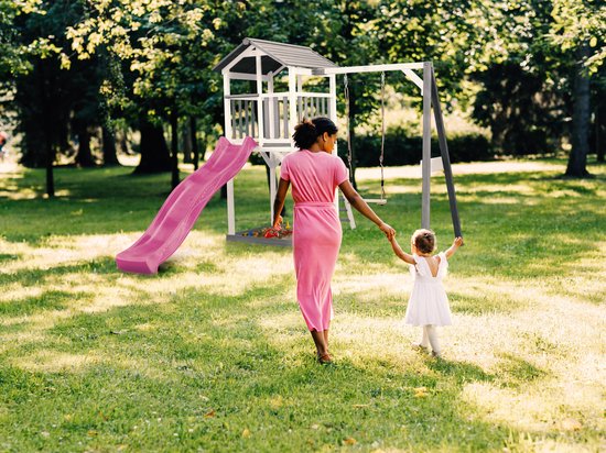 Moeder in roze jurk houdt haar dochter vast bij een speeltoren met paarse glijbaan en schommel in een zonnige tuin.